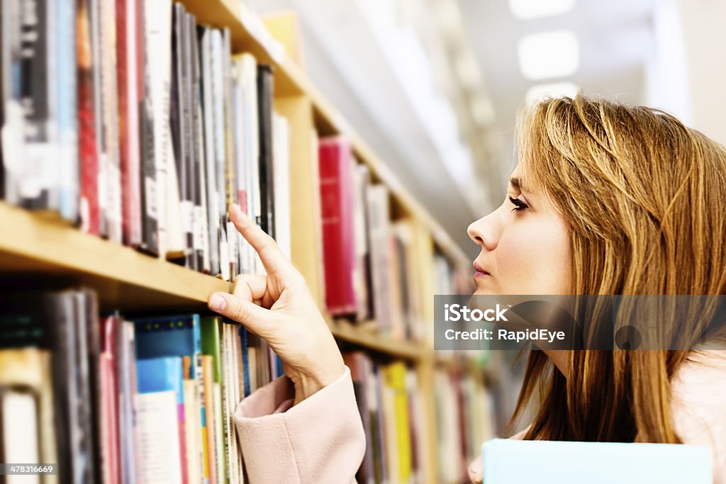 Linda mulher loira pesquisas as estantes de livros em uma biblioteca - Foto de stock de 20 Anos royalty-free