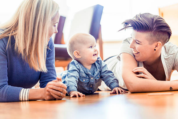 Smiling lesbians with baby lying on hardwood floor Smiling lesbian couple with baby lying on hardwood floor. Homosexual partners with toddler in living room. Happy family of three at home. unknown gender stock pictures, royalty-free photos & images