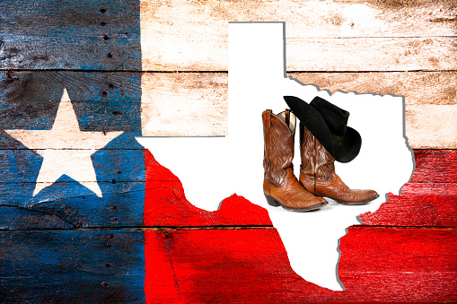 Rustic Texas, USA flag created from old wooden boards that have been painted red, white and blue with a state of Texas map outline placed inside the flag.  Cowboy boots and hat inside the state cutout.  A lone star to left. Weathered, handmade craft. Unique background. White map outline. 