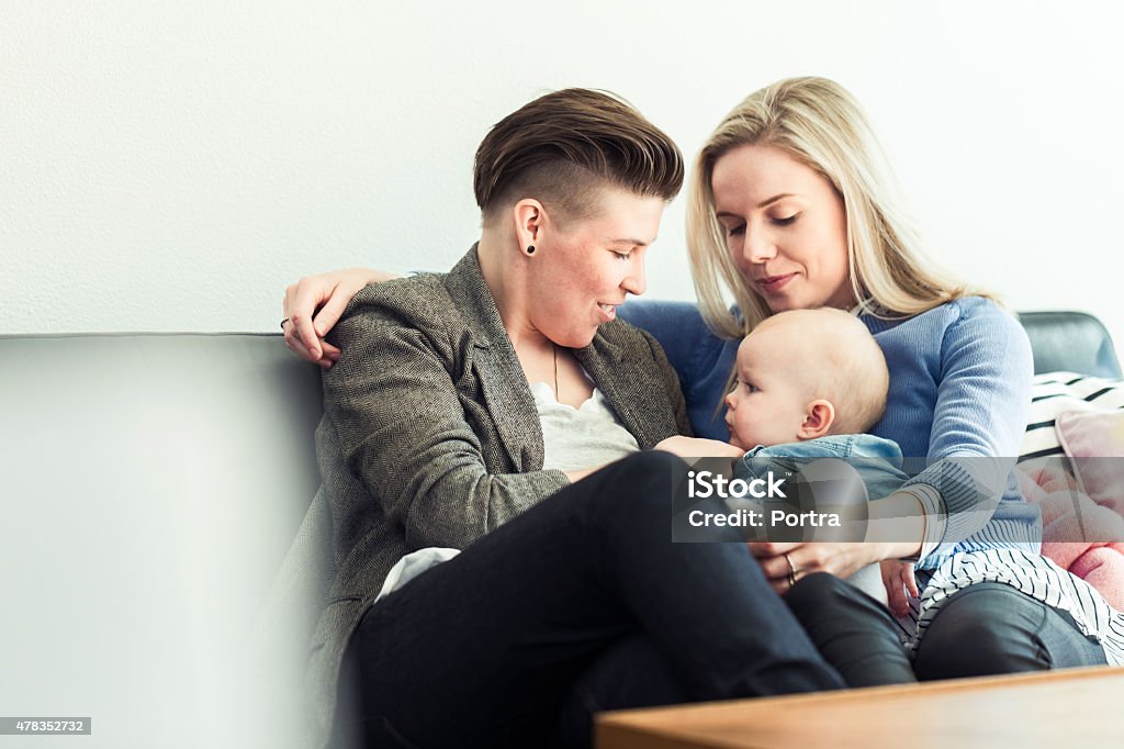 Young lesbian couple playing with baby on sofa Young lesbian couple playing with baby on sofa. Loving female partners with cute toddler in living room. Happy family of three. Gay Couple Stock Photo
