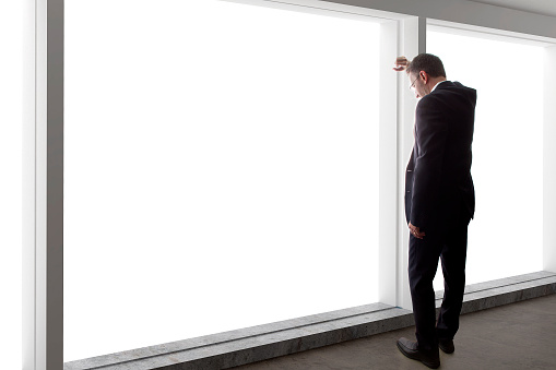 Middle aged businessman looking out a bright office window and thinking.  The entrepreneur is middle aged and wearing a suit.  He looks like he is a boss or CEO.  He is in an office loft with big windows.