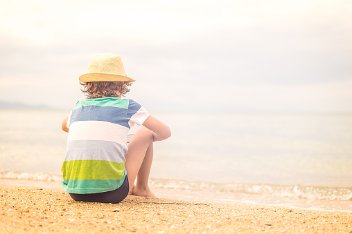 Little boy with straw hat sitting at the beach and looking away