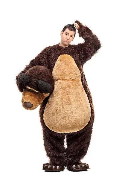 Full length portrait of a confused young man in a bear costume scratching his head and looking at the camera isolated on white background