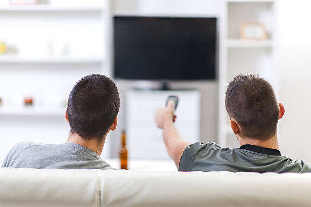 Rear view of two men watching TV at home. Rear view of two male friends sitting on the sofa at home and watching TV. One of them is holding remote control and changing channels. couch back stock pictures, royalty-free photos & images