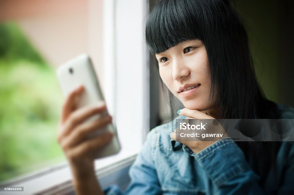 Asian teenager girl using mobile phone while sitting near window. Indoor close-up shot of Asian teenager girl using mobile phone while sitting near window. She is in blue denim shirt looking at mobile phone. The Horizontal head and shoulders composition with copy space was shot at day time in natural available light using wide open aperture f/1.8. Chin Stock Photo