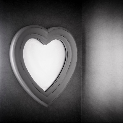 Monochrome image of a steamed-up, heart-shaped bathroom mirror, with light streaming in from a window to the left. Dreamy, soft image with very shallow focus.