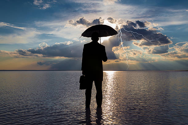 Businessman in trouble concept A rear view of a businessman in black suit with a briefcase and a black umbrella standing at water edge and looking rainstorm clouds at sunset. Lightning is seen descending from a gray and cloudy sky. recession protection stock pictures, royalty-free photos & images