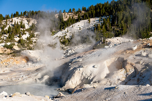 A landscape of Lassen Volcanic National Park, California. Horizontal image shows the steamy hot springs, surrounded by trees on a mountain side.