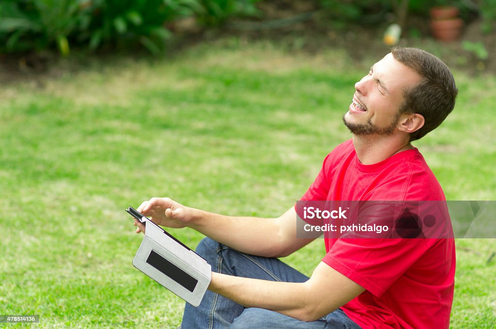 Man studying in park Man wearing jeans and red t-shirt sitting on grass with tablet laughing 2015 Stock Photo