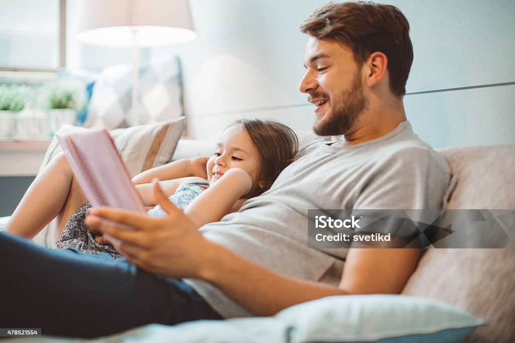 Father and daughter enjoying at home. Father and daughter enjoying at home. Sitting on bed and reading book together. 2015 Stock Photo