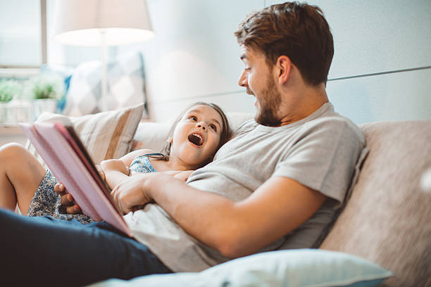 Father and daughter enjoying at home. Father and daughter enjoying at home. Sitting on bed and reading book together. family with one child stock pictures, royalty-free photos & images