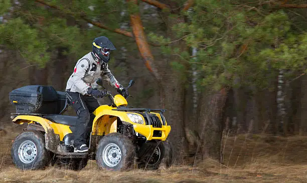Horizontal motion portrait of a man in safety helmet and goggles driving mud-covered yellow ATV 4x4 quad bike in gray sport jacket in the woods.