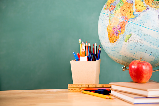 It's back to school time!  A globe with school supplies on top of school desk with chalkboard in background. Elementary school supplies include: apple, textbooks, pencils, pens, crayons, ruler, and more.  The blank blackboard in the background makes perfect copyspace!  Education themes. 