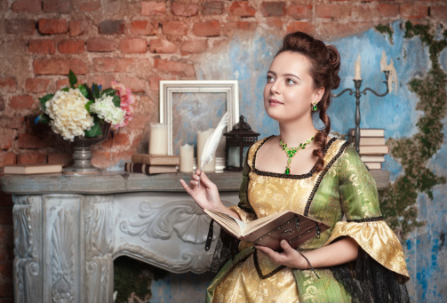 Beautiful young  woman in long medieval dress writing in diary near fireplace