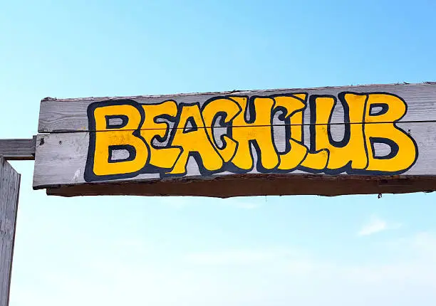 Beach club signboard with yellow letters on a wooden plank. Entrance sign of a beach restaurant or Beachclub. Close-up with blue sky.