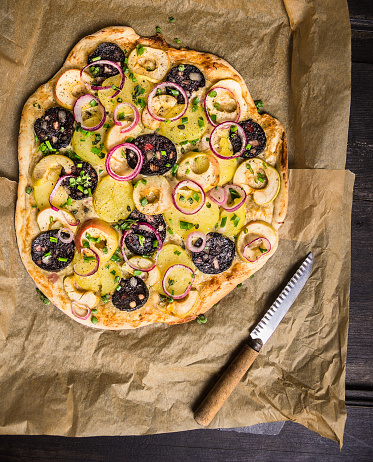 tarte flambee with potato, red onion,appels and  blood sausage on baking paper with vintage knife, top view