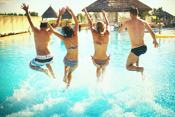 Group of young adults jumping in swimming pool with their arms raised.There are two girls and two guys. They are on vacation and enjoying their hotel. Rear view.