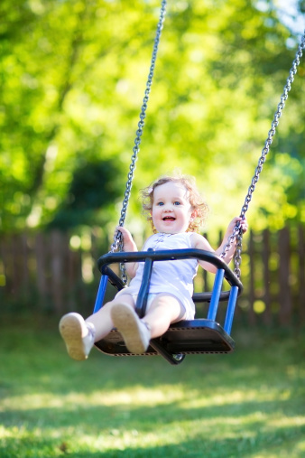 Adorable baby girl with big beautiful eyes and curly hair having fun at a swing on a playground in a sunny summer park