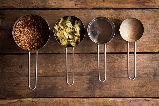 An overhead shot of a collection of basic home brew beer ingredients, sitting on a rustic wooden background, lit by natural daylight. 