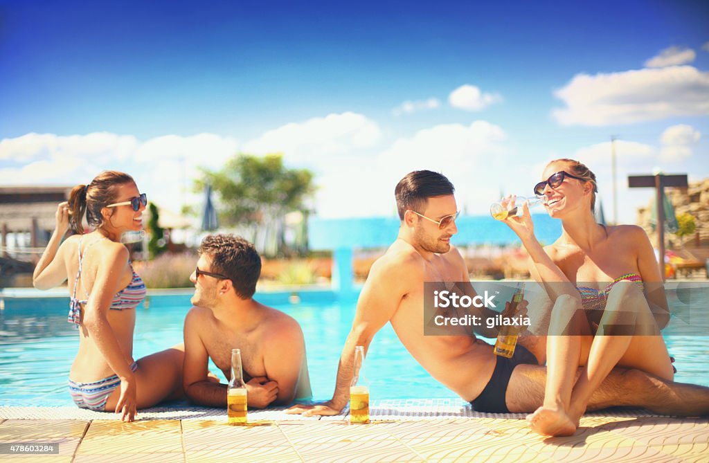 People enjoying drinks by the swimming pool. Two couples having beers while relaxing by the poolside of the hotel they stayed in. Their vacation has just started. Mid 20's men and women Each person is wearing sunglasses. Tourist Resort Stock Photo