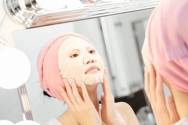 A Japanese woman puts on a facial mask treatment as she looks at her reflection in the mirror.