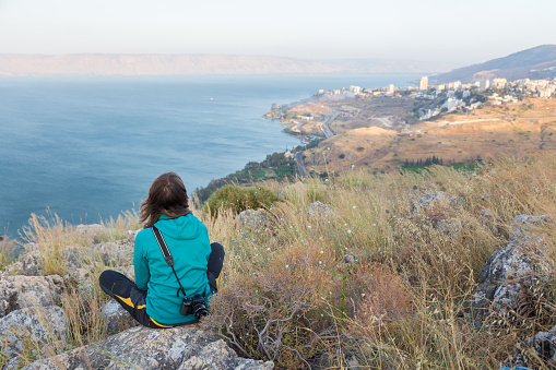 Young photographer woman sitting resting mountain rocks above city town Tiberias and Galilee Kineret sea lake, watching Golan Heights range mountains. Rear back view. Israel.
