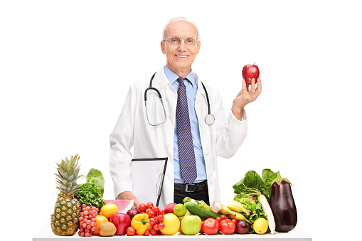 Mature doctor holding an apple and standing behind a table full of fruits and vegetables isolated on white background