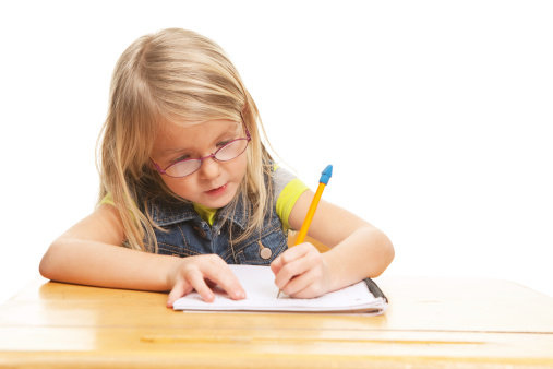 A six year old girl, seated at a school desk, is writing in a note book with a pencil. The desk has some wear and scratches.