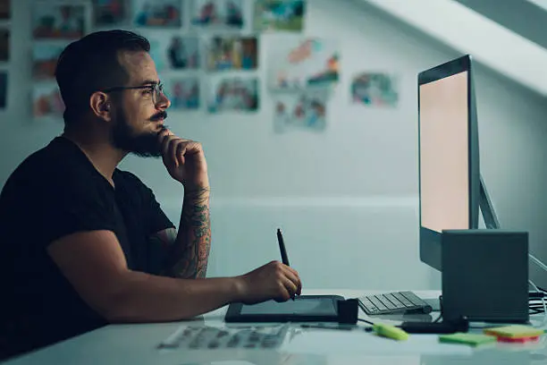 Graphic Illustrator or designer working in his own office. Using digitized graphic tablet, drawing with digitized pen. Working late. Side view. White screen so you can add your own project on monitor.