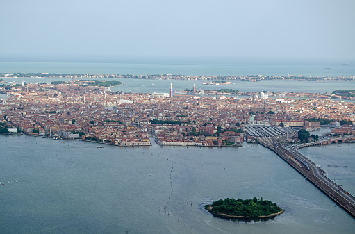 View from above of the city of Venice, Italy.  A train is crossing the causeway to the mainland in the distance large ships are anchored before entering the lagoon.  Spring day in May.