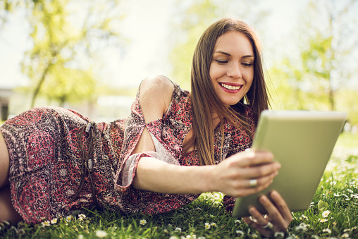Young smiling woman enjoying in grass during spring day and using touchpad.