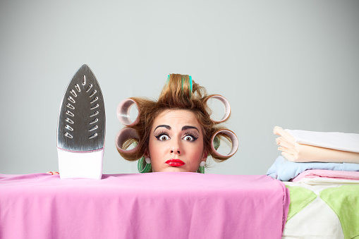 Studio portrait of funny stereotypical housewife hiding behind ironing board. Nerdy wife with curlers in hair is making confused and surprised face. Behind her is gray background. She is looking to camera.