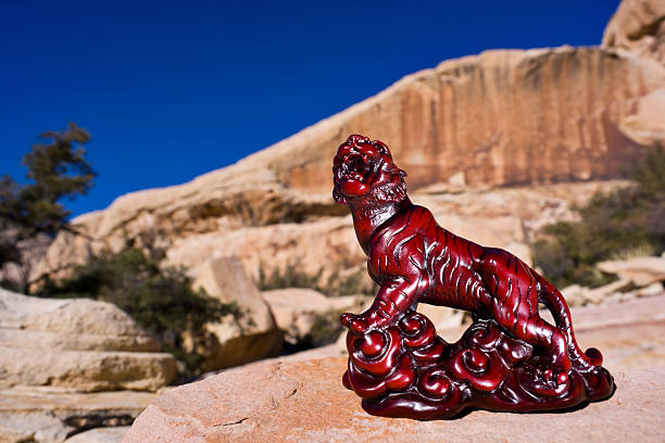 A small, red, wooden, carved statue of a Chinese tiger (one of the Chinese Zodiak animals).  The figurine sits on sandstone rocks, with sandstone hills, desert plants and a blue sky in the background.  The image is said to bring good luck.