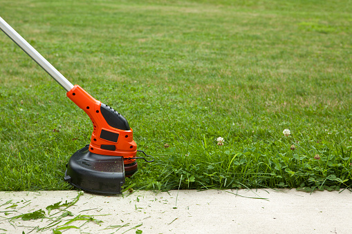 Close-up of a string weed trimmer trimming the grass along a concrete sidewalk.