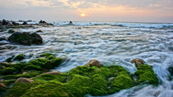 Beautiful, amazing landscape of nature with green moss cover stone, the sun go up at horizon, very white waves surf onto large rock make mysterious, peaceful, impressive scene like paradise on sea