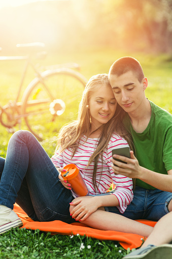 Teenage couple holding each other and looking something on a cell phone. The boy is wearing green t-shirt and the girl is dressed in striped shirt and jeans. In the background is a bicycle and spring meadow bathed in evening spring sunlight