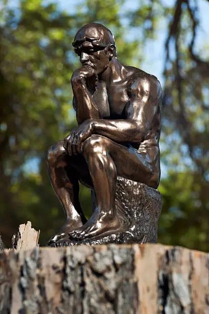 View of a small bronze replica of Rodin's The Thinker statue, sitting in a spot of light in a forest.  The Thinker is often used to represent philiosphy.