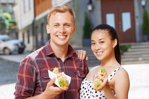 Going to eat. Young Caucasian man and Asian woman standing on street and holding hotdogs
