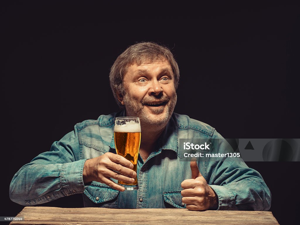 The smiling man in denim shirt with glass of beer Enjoying his favorite beer.  The front view of handsome smiling  man as fan in denim shirt with glass of beer, sitting at the wooden table 2015 Stock Photo