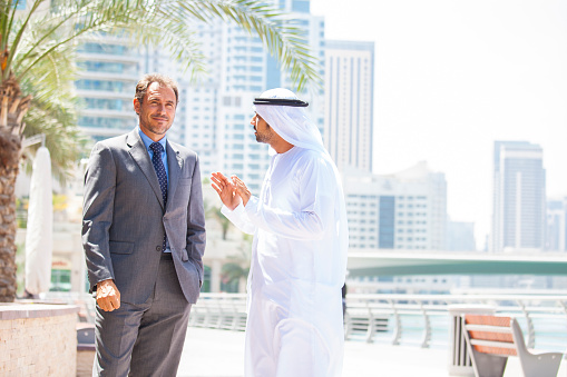 Businessmen standing in front of Dubai Marina skyline. They are talking one each other. Arab businessman is  worn in traditional male dress  kandura (dishdasha) with  kaffiyeh (white headdress of cotton fabric) held in place by a rope (agal). Concept for modern business. Image is taken during Dubai istockalypse.