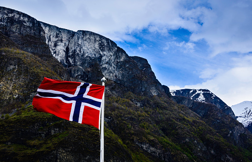 Norwegian flag flies against a backdrop of Aurlandsfjord, a tributary of the giant Sognefjord. The fjord is know for steep mountainsides, waterfalls, snow fields, and picturesque farms and hamlets.