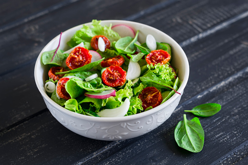 salad with fresh vegetables, garden herbs and sun-dried tomatoes in a white bowl on a dark wooden background