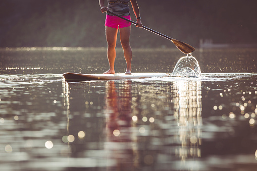 Young Asian female paddle boarding in calm waters during sunset.  Focus on water formation and pattern created by oar.