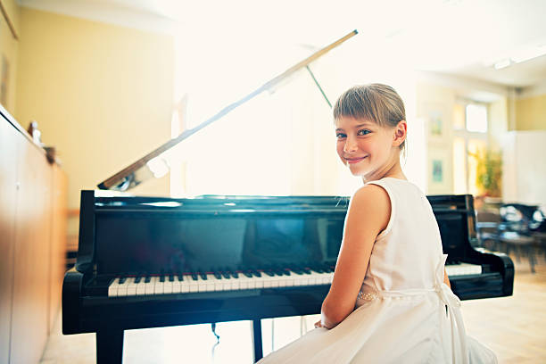 Little girl playing on grand piano Little girl playing grand piano in a sunny class room. She is preparing for a piano performance in music school. The girl aged 9 is sitting at the grand piano, looking over shoulder and smiling to the camera. The girl is wearing white dress. girl playing piano stock pictures, royalty-free photos & images