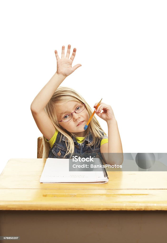 Child Girl Sitting at School Desk Raising Hand A six year old girl, seated at a school desk, is writing raising her hand to ask or answer a question. The desk has some wear and scratches. Child Stock Photo