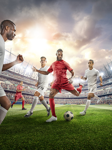 A male soccer player makes a dramatic play with a ball.  The opposite team players are going to block the ball. All players are wearing generic unbranded soccer uniform. A wide angle panoramic image of a outdoor soccer stadium outdoor stadium or arena full of spectators under a dramatic stormy evening sky at sunset. The image has depth of field with the focus on the foreground part of the pitch with intentional lensflare