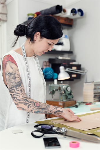 Portrait of a professional dressmaker at work in her design studio. She is carefully marking out the pattern from a paper template  onto the material with a piece of  chalk. Colour, vertical with a very shallow point of focus on the model's face, she is wearing a white sleeveless top that shows of her full sleeve tattoo she is half Japanese half european with dark hair.