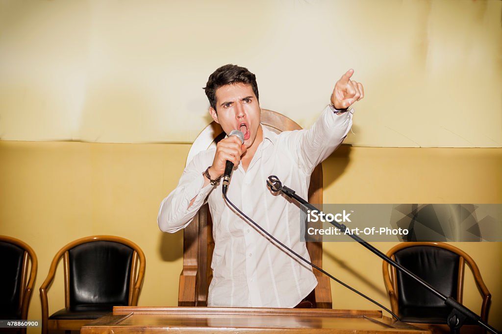 Passionate Man Speaking into Microphone in Meeting Determined Young Man Speaking Passionately into Microphone and Pointing into Air, Rallying Support for Political Cause in Meeting Desk Stock Photo