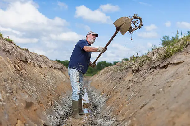 A bearded, older workman busy digging a huge ditch across a field using only a long-handled shovel.