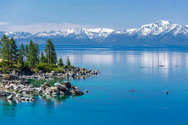 Picture of Lake Tahoe from east shore. There is a snow on the Sierra Nevada Mountains and some white clouds with reflection in turquoise waters of the lake. There is a paddle boarder arriving to the beach. Sand Harbor State Park is visible on the left side of the photograph.